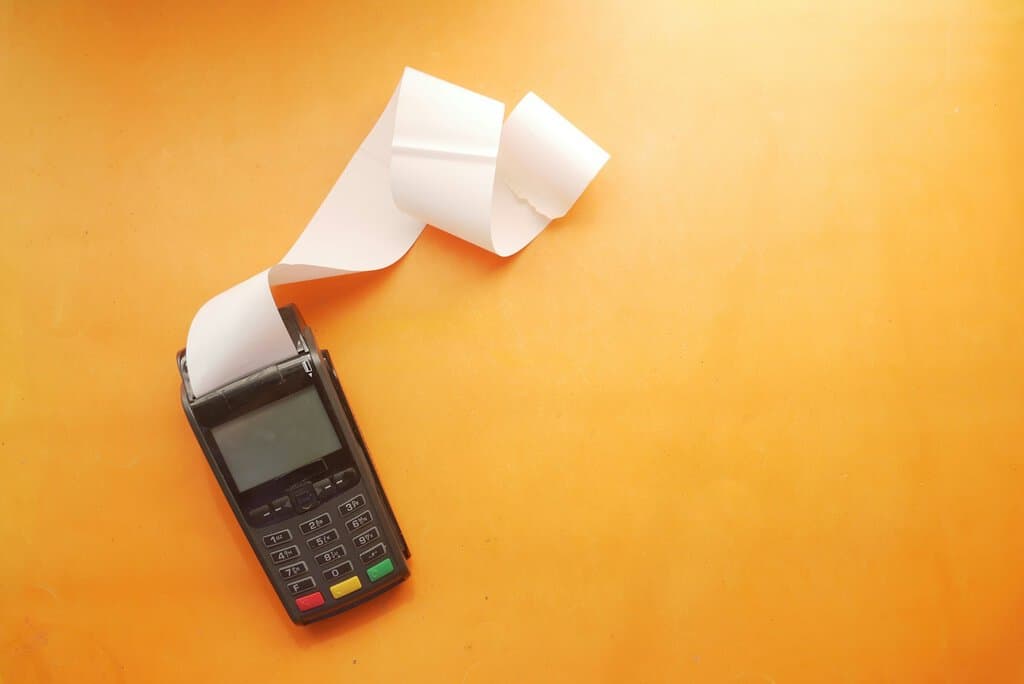 A black payment terminal with a long white paper receipt on orange background
